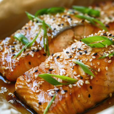 Close-up of glazed salmon fillets topped with sesame seeds and sliced green onions, set in a savory sauce. The fish appears tender and perfectly cooked, garnished for an appetizing presentation.