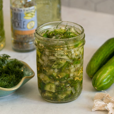 A glass jar filled with sliced cucumbers, fresh dill, and pickling liquid sits on a countertop. Whole cucumbers, a bowl of dill, and a jar of dill pickle seasoning are nearby.