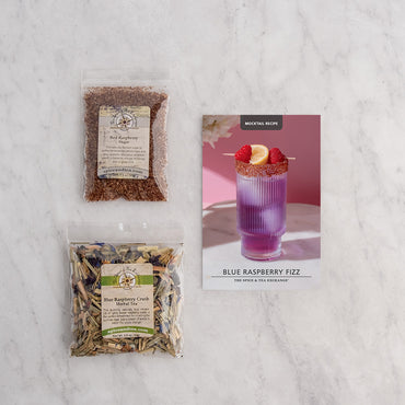 The Blue Raspberry Fizz (Kit) features two spice packetsāRaspberry Sugar and Blue Raspberry Crushāarranged on a light surface next to a recipe card showing a vibrant mocktail with raspberries and a sugared rim.