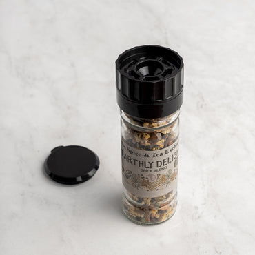 A glass jar of Earthly Delight Spice Blend with a black grinder top sits on light marble, ready to spice up meats, stews, and soups. The jarās black cap rests nearby.