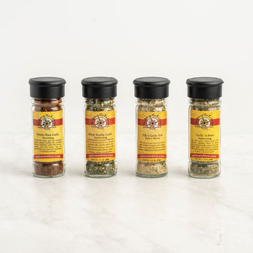 Four spice jars with black lids are lined up on a white surface. The Garlic Lover's 4-Pack Gift Box includes: Smoky Black Garlic, Black Truffle Garlic, Viki’s Garlic Fix, and Garlic n Herb.