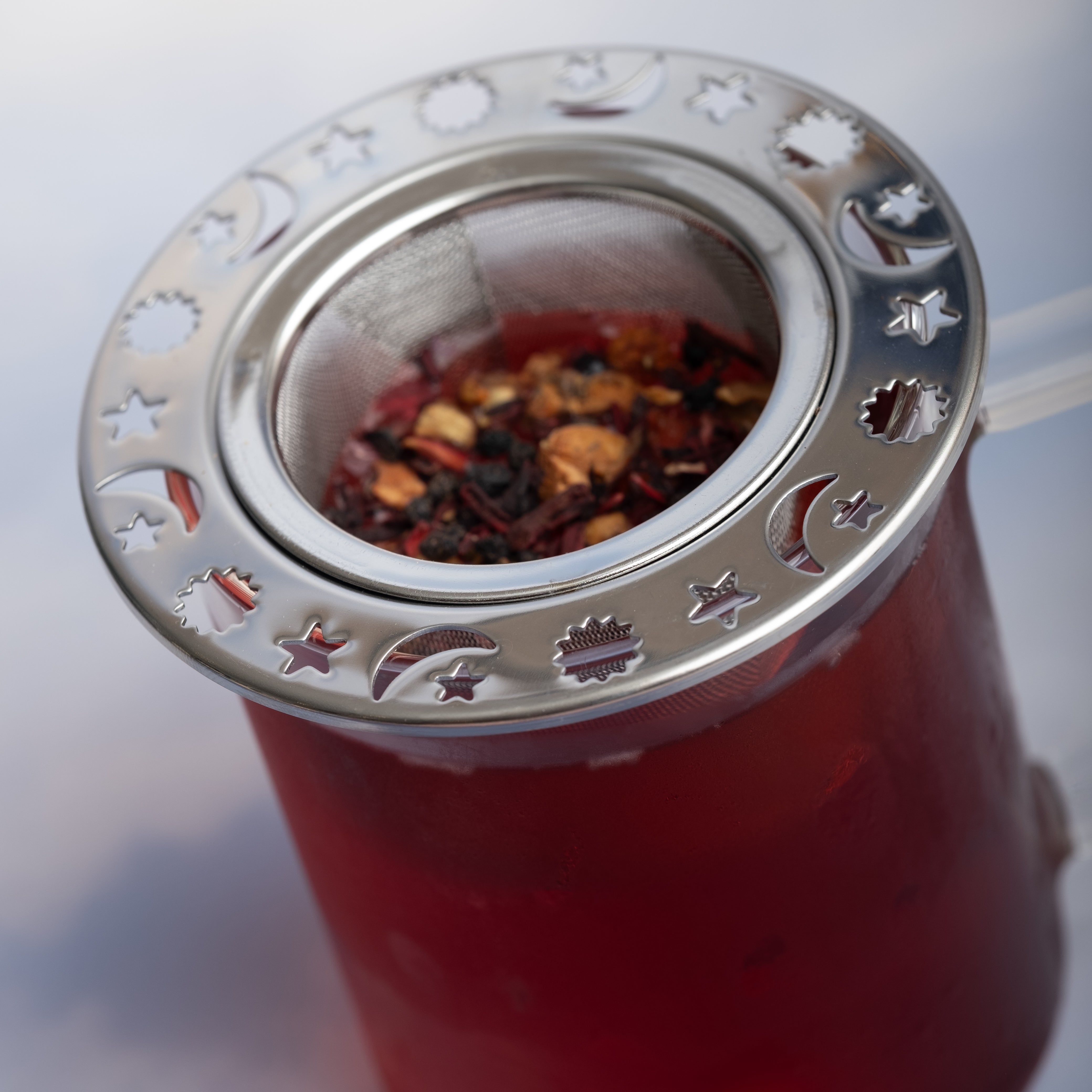 A glass mug of red herbal tea brewed with the Moon and Stars Infuser, featuring celestial cut-outs, holding aromatic loose-leaf tea and fruit pieces.