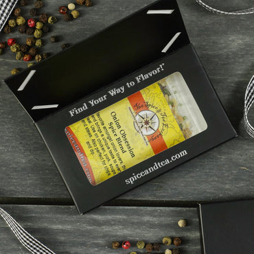A box labeled "Find Your Way to Flavor!" holds a jar of Onion Obsession Spice Blend - Barter Box from The Spice & Tea Exchange—an ideal gift. Peppercorns and ribbon decorate the dark wooden surface around it.