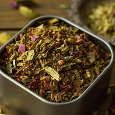 A close-up of a metal container filled with Beauty Rest Herbal Tea, a colorful blend with relaxing botanicals like fennel seeds, cardamom pods, dried rose petals, and valerian root. A small dish of extra spices appears in the background.