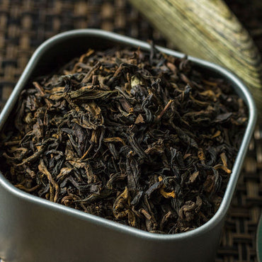 A close-up of Organic Black Pu-erh Tea from Yunnan fills a square metal container on a woven brown surface. The dark, twisted leaves suggest their signature earthy flavor.