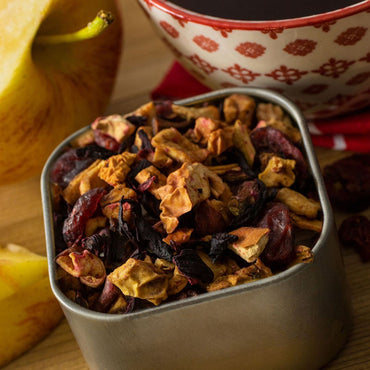A close-up of a metal container filled with Cranberry Apple Tart Herbal Tea and dried fruit, beside a sliced apple and a patterned bowl of dark tea on a wooden surface.