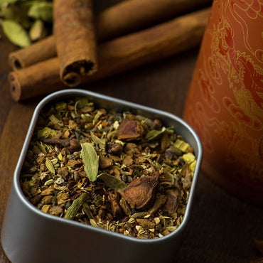 A close-up of Rooibos Chai Herbal Tea with cardamom and ginger in a square metal tin, cinnamon sticks nearby, and a decorative container in the background on wood, showcasing its bold flavor.