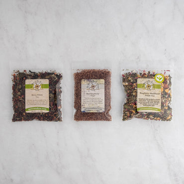 Three clear bags of loose leaf tea labeled Berry White Tea, Red Raspberry Sugar, and Raspberry Shortbread White Tea from the Raspberry Shortbread Sampler are displayed side by side on a light marble surface.