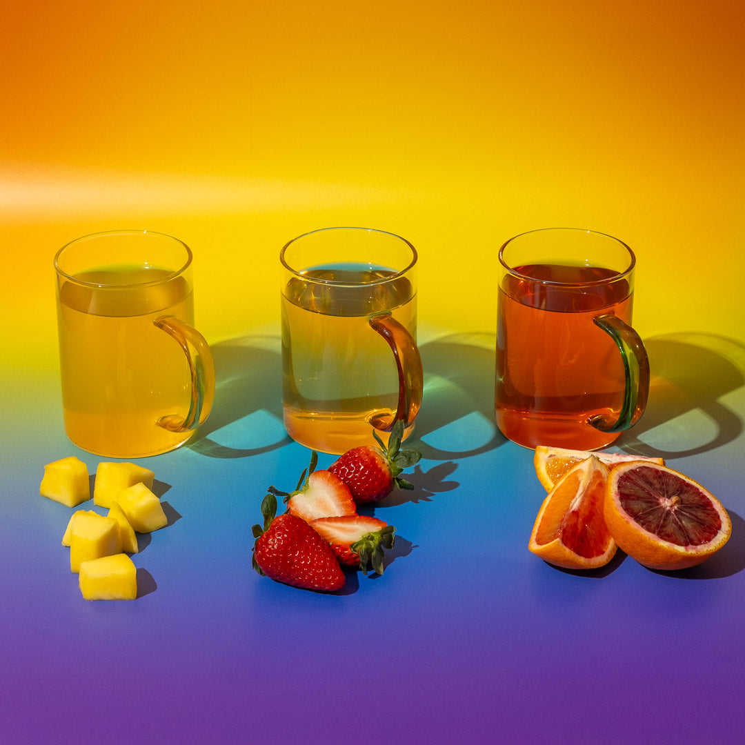 Steep the Rainbow Sampler showcases three glass mugs of herbal and fruity teas on a yellow, orange, and blue background, with diced mango, sliced strawberries, and blood oranges in front for a vibrant tea tasting experience.