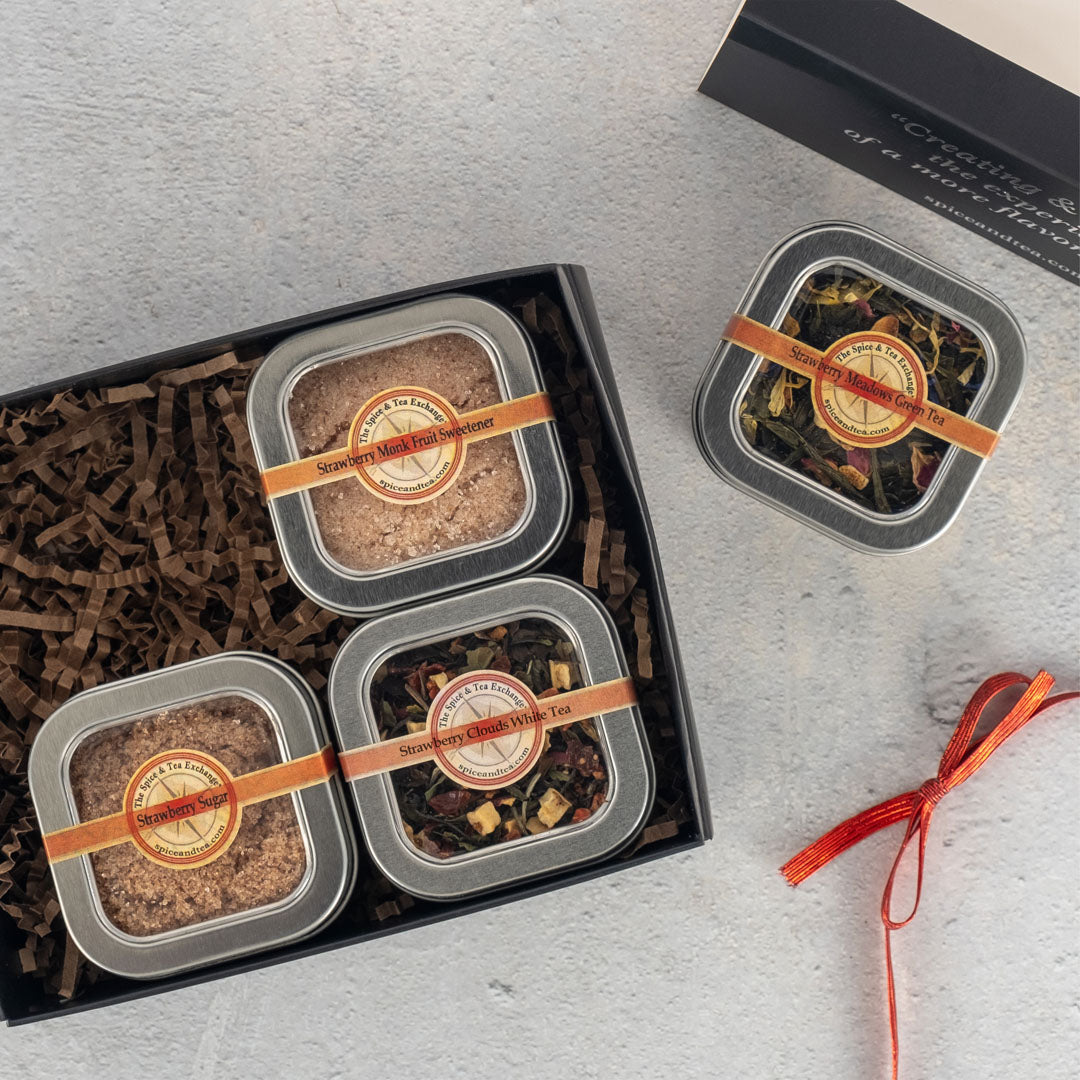 The Strawberry Fields 4-Tin Gift Box includes three metal tins of loose leaf tea and a strawberry sweetener powder, all arranged on brown shredded paper, with an extra tin and a red ribbon displayed beside the box on a light grey surface.