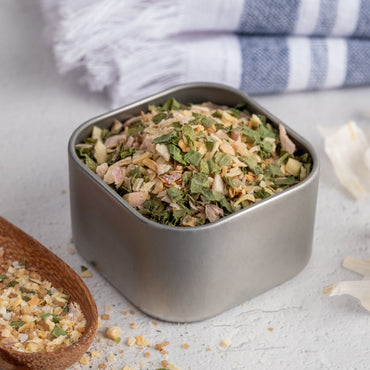 A square metal container of Vik's Garlic Fix! Spice Blend rests on a light surface, with a wooden spoon holding some of the blend nearby and a striped cloth in the background.