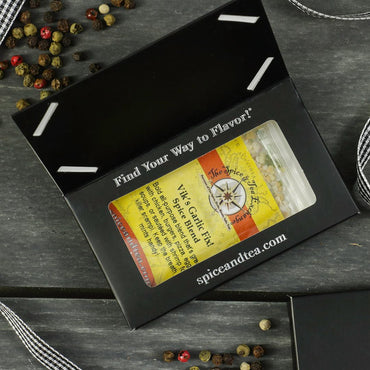 A black gift box with a clear window displays the Vik's Garlic Fix Spice Blend - Barter Box. Peppercorns, ribbon, and a business card are arranged on a gray wooden surface around the gift sampler.