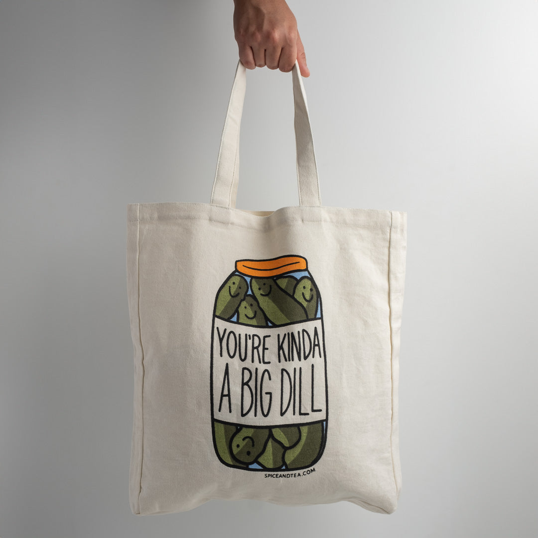 A hand holds The Spice & Tea Exchange "You're Kinda A Big Dill Tote Bag," which features smiling pickles in a jar with the text “You’re Kinda a Big Dill” on a plain light gray background.