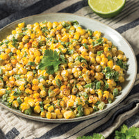 A plate of Mexican street corn salad garnished with cilantro, featuring roasted corn, chopped green onions, and spices, with a lime half in the background on a striped cloth.