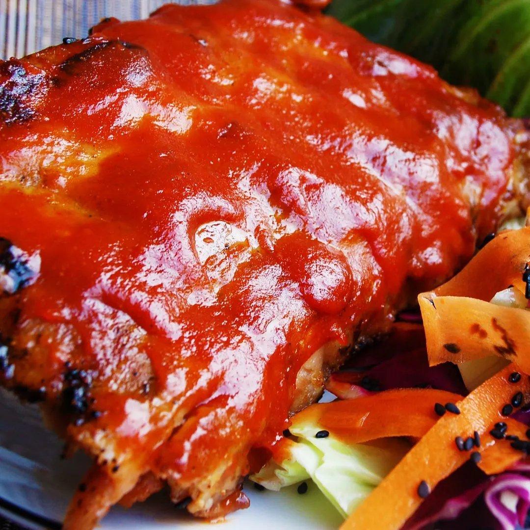 Close-up of barbecued ribs covered in a shiny red sauce, served with a side of sliced carrots, cabbage, and black sesame seeds.