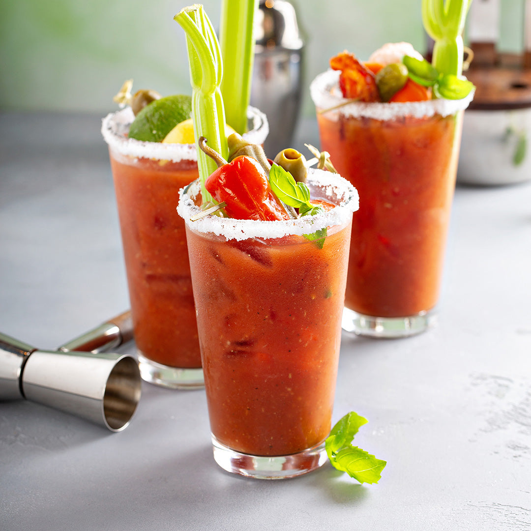Three Bloody Mary cocktails in salt-rimmed glasses, garnished with celery stalks, lemon and lime wedges, olives, and pickled vegetables, sit on a light gray surface next to a jigger and cocktail shaker.