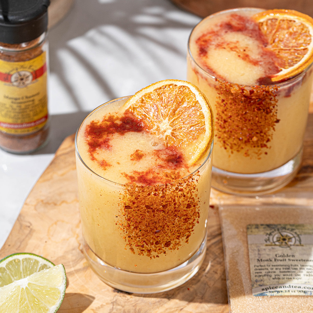 Two glasses of a creamy orange beverage garnished with a dried orange slice and chili powder on the rim, surrounded by a cut lime, spice jar, and sweetener packet on a wooden board.