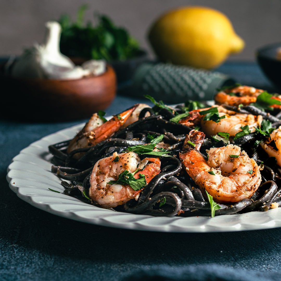 Squid Ink Linguine with Shrimp (Kit): White plate of black linguine topped with grilled shrimp and parsley. Lemon, garlic, and a wooden bowl of herbs on a dark blue surface complete this perfect meal for shrimp pasta lovers.