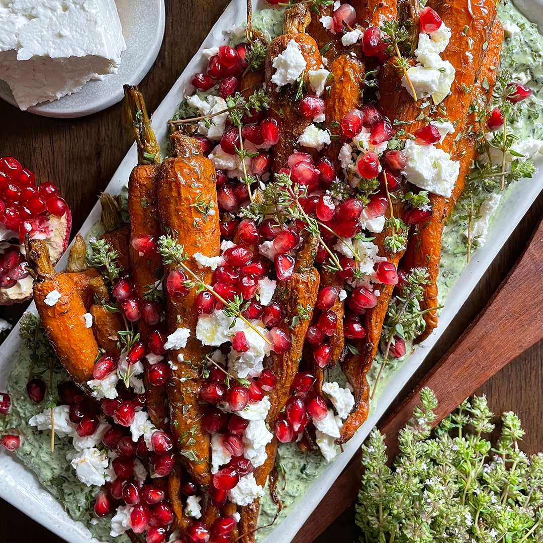 Roasted Carrots (Kit) features roasted carrots with crumbled feta, fresh herbs, and pomegranate seeds on creamy Green Goddess dip, served on a rectangular white platter.