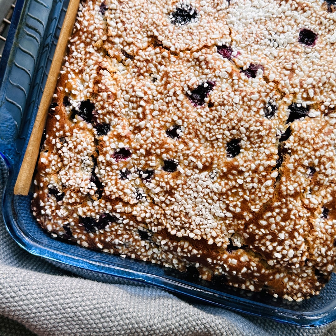 A close-up of the Cardamom Pearl Tea Cake (Kit) in a glass dish, topped with Swedish pearl sugar and dark berries, resting on a woven cloth.