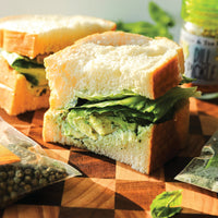 The Green Goddess Sandwich (Kit), a thick vegetarian lunch sandwich with lettuce and creamy green spread, sits on a wooden board beside Green Goddess Seasoning, Dill Pickle Sea Salt packets, and a Dill Pickle jar in bright natural light.