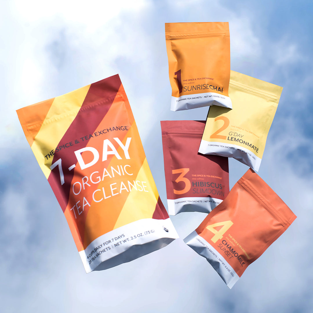 Five colorful wellness tea packets labeled 7 DAY ORGANIC TEA CLEANSE float against a blue sky. Each numbered packet features unique organic teas: Sunrise Chai, Lemonmate, Hibiscus Slimdown, and Chamomile Unwind.