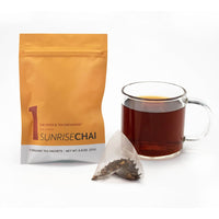 A glass mug of dark tea sits beside a yellow-orange 7 Day Organic Tea Cleanse sachet and a pyramid tea bag filled with loose leaves—an inviting start to your daily organic tea cleanse, set against a white background.
