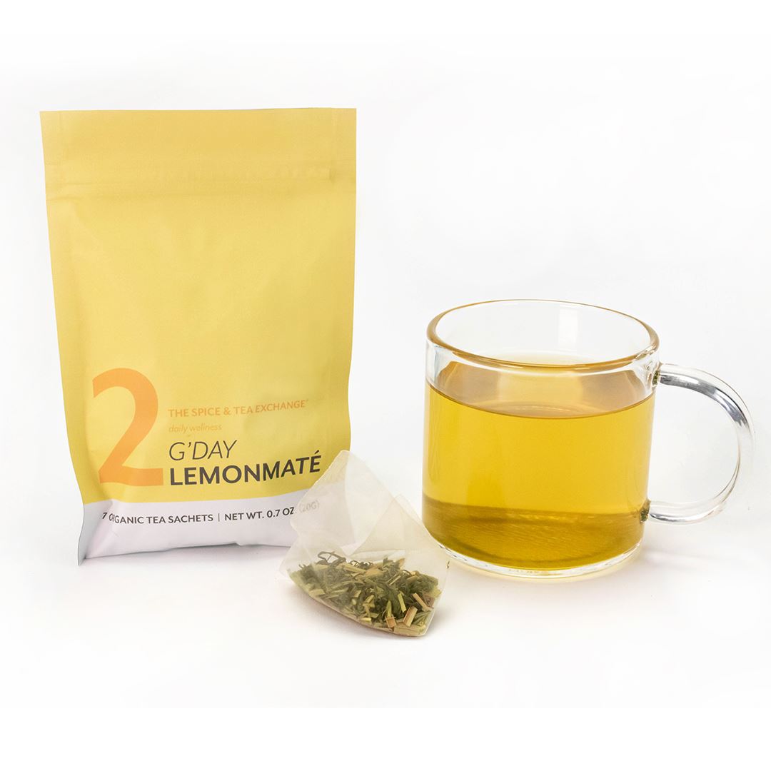 A clear glass cup of yellow tea sits beside a yellow package labeled 7 Day Organic Tea Cleanse and a pyramid-shaped tea bag with visible loose leaves, perfect for those seeking a refreshing wellness tea experience.