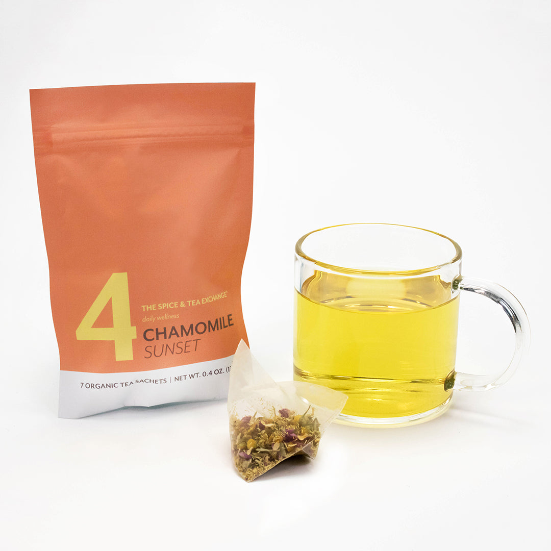 A glass cup of yellow chamomile tea sits next to a pyramid herbal sachet and a peach packet labeled 4 Chamomile Sunset, providing a soothing option in the 7 Day Organic Tea Cleanse for your wellness routine.