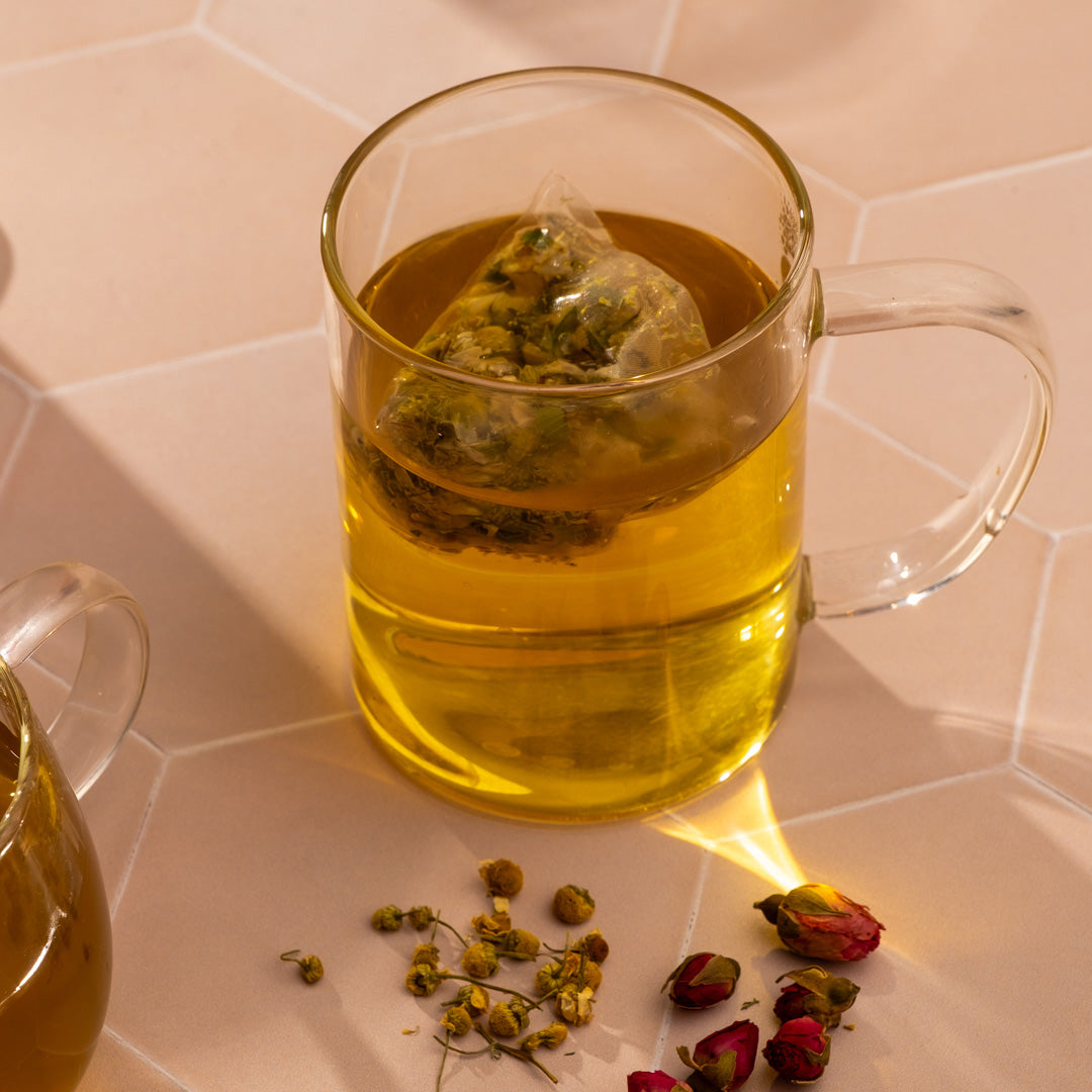 Clear glass mug with yellow tea and a tea bag on a tiled surface with dried herbs.