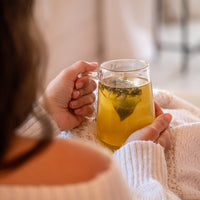 A person in a cozy white sweater holds a clear glass mug of hot tea, herbs visible inside, with a softly blurred background creating a warm, relaxing atmosphere—perfect for enjoying the 7 Day Organic Tea Cleanse.