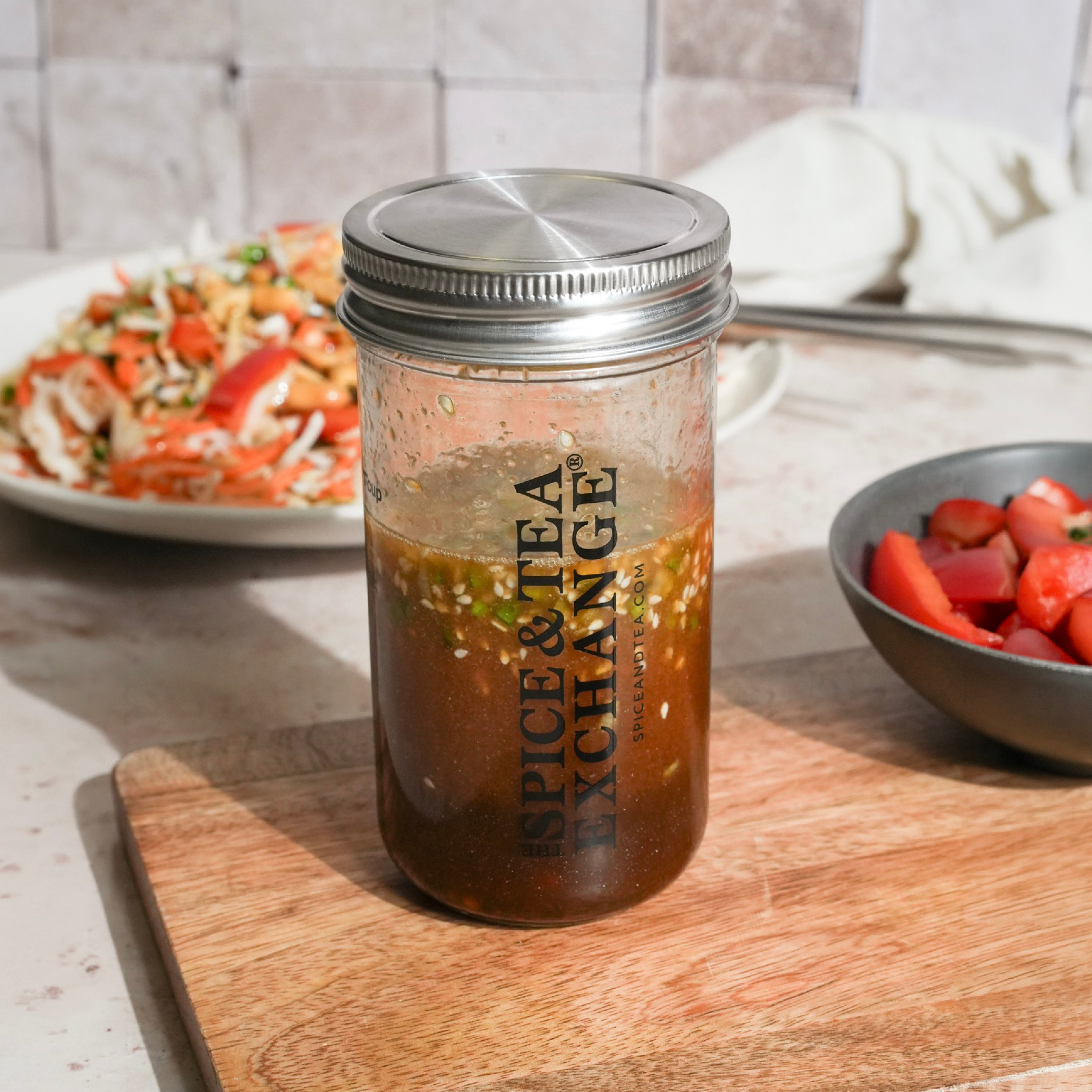 A jar labeled "Asian Dressing (Kit)" from The Spice & Tea Exchange sits on a cutting board, accompanied by a bowl of chopped strawberries and a plate of tossed salad in the background.