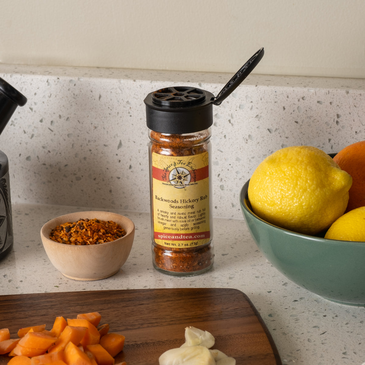 A jar of Backwoods Hickory Rub Seasoning sits on a kitchen counter beside lemons, oranges, a wooden bowl of spices, chopped carrots, and garlic cloves.