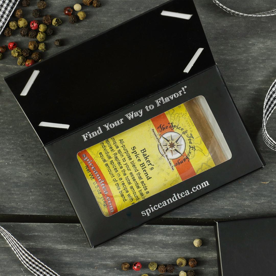 A black box with a window displays the Baker's Spice Blend - Barter Box, an ideal gift for baking enthusiasts. Decorative peppercorns and ribbon are scattered on a dark wooden surface.