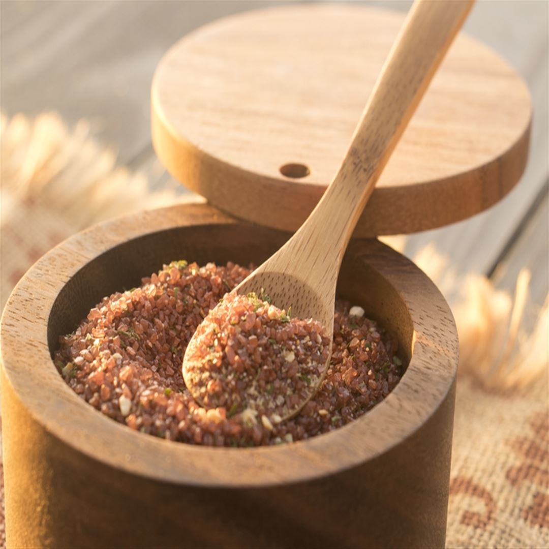 The Bamboo Salt Spoon Mini rests inside a wooden container filled with coarse reddish-brown seasoning, its open lid revealing the texture and sunlight highlighting its stylish appeal for your salt accessories collection.