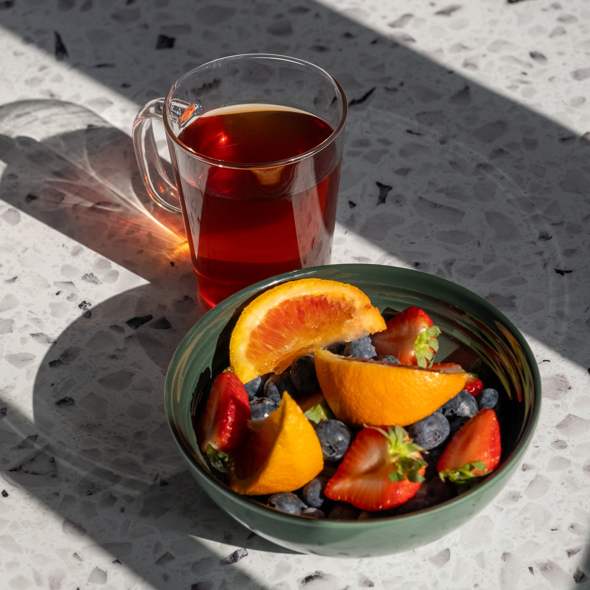 A glass mug of Berry Bouquet Herbal Tea and a green bowl with orange slices, strawberries, and blueberries sit on a terrazzo surface in sunlight, casting distinct shadows.