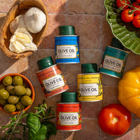 Five tins of Black Truffle Extra Virgin Olive Oil - Mini are displayed on tiles with fresh garlic, olives, yellow bell pepper, and red tomatoes, bathed in sunlight that casts natural shadows.