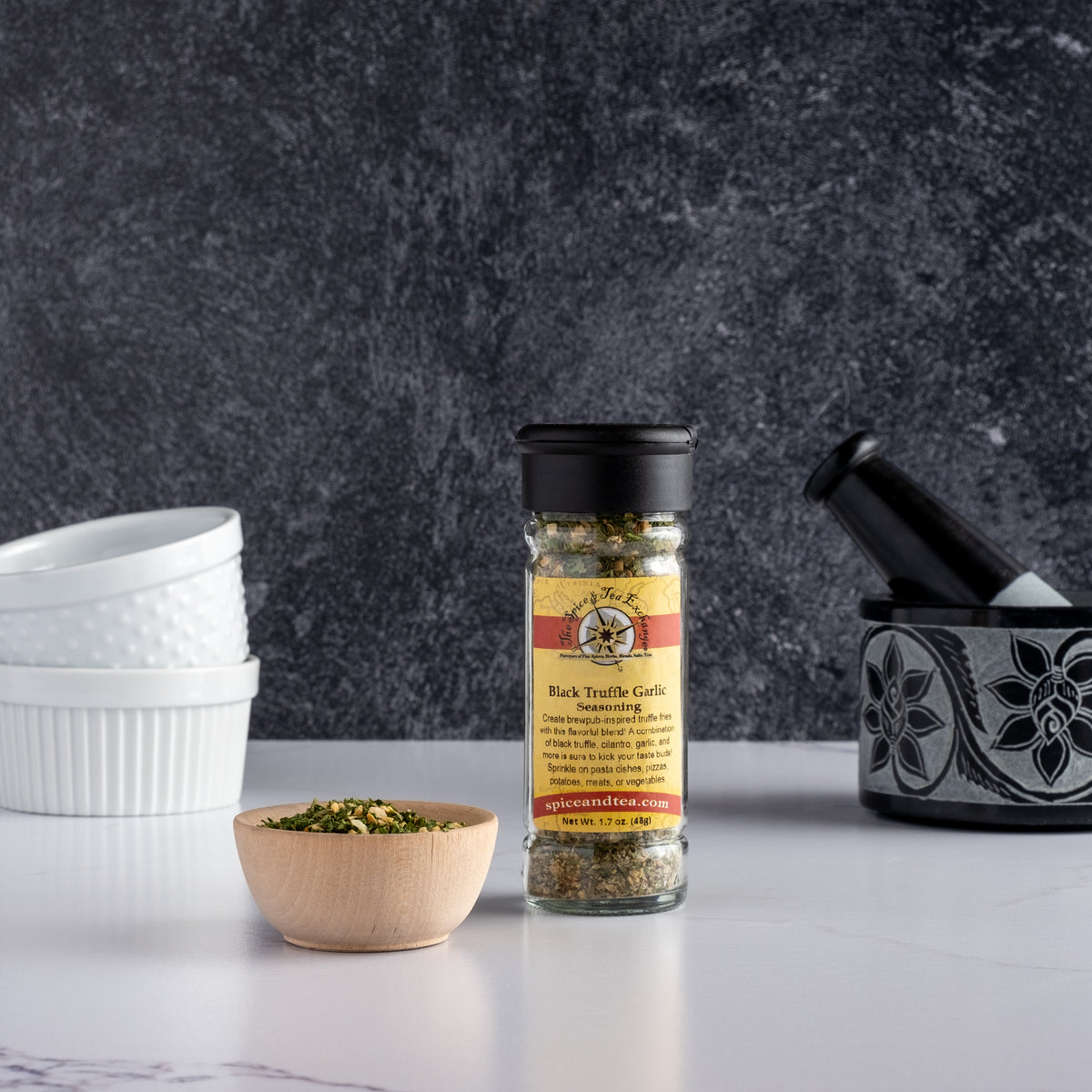 A jar of Black Truffle Garlic Seasoning sits on a white surface next to a wooden bowl of seasoning, white ramekins, and a black mortar and pestle, all arranged against a dark textured background.