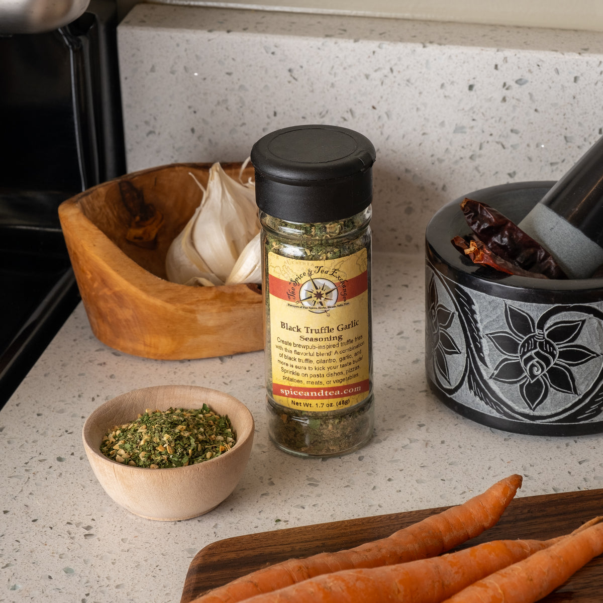A jar of Black Truffle Garlic Seasoning sits on a kitchen counter beside garlic cloves, a mortar and pestle with dried chilies, a bowl of seasoning, and whole carrots—just what you need to upgrade your truffle fries.