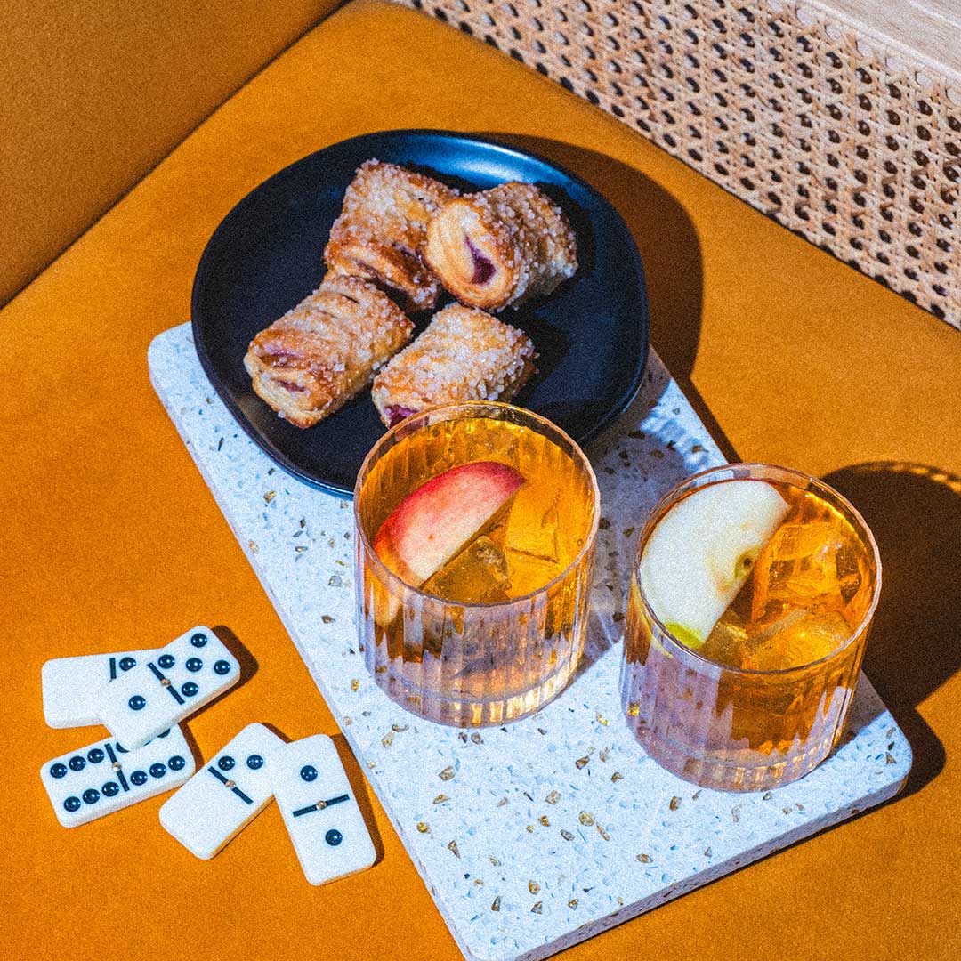 Two glasses of a beverage with apple slices, a plate of pastries, and dominoes on a textured surface.