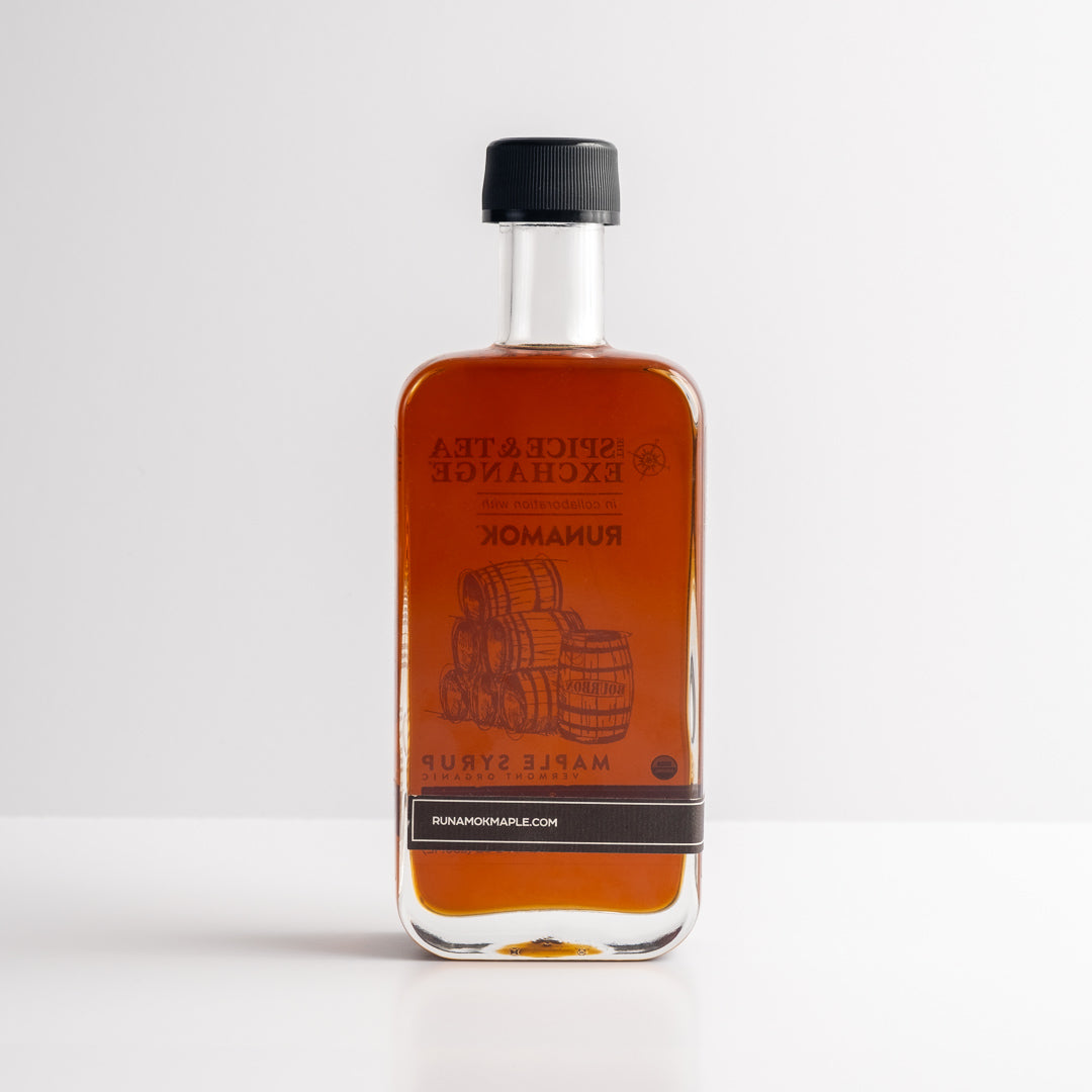 A rectangular glass bottle of Organic Barrel-Aged Bourbon Maple Syrup with dark amber syrup stands upright on a white surface. Its label features barrels and "pure Vermont maple syrup," finished with a black cap.