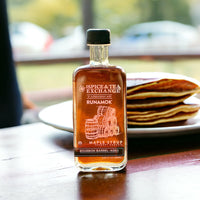 A bottle of Organic Barrel-Aged Bourbon Maple Syrup from Spice & Tea Exchange in collaboration with Runamok sits on a table before a plate of pancakes, the softly blurred background highlighting its rich color.