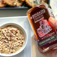 A hand holds a bottle of Organic Barrel-Aged Bourbon Maple Syrup above a bowl of oatmeal topped with syrup and spices. The bottle is labeled "Spice & Tea Exchange in collaboration with Runamok.