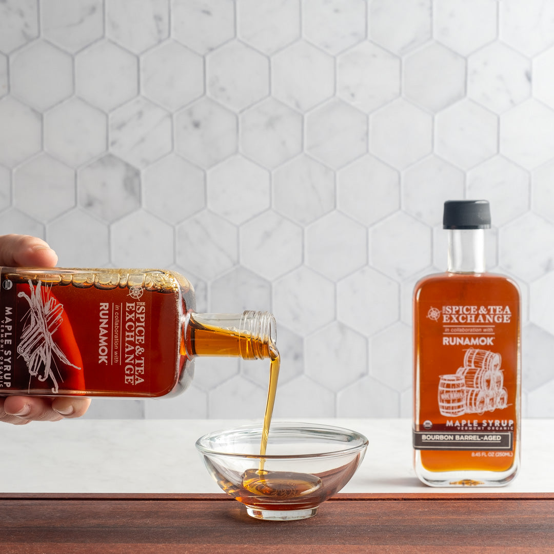 A hand pours Organic Barrel-Aged Bourbon Maple Syrup from a glass bottle into a small bowl, with another bottle of the syrup standing upright on a marble countertop in front of a hexagonal tile backsplash.