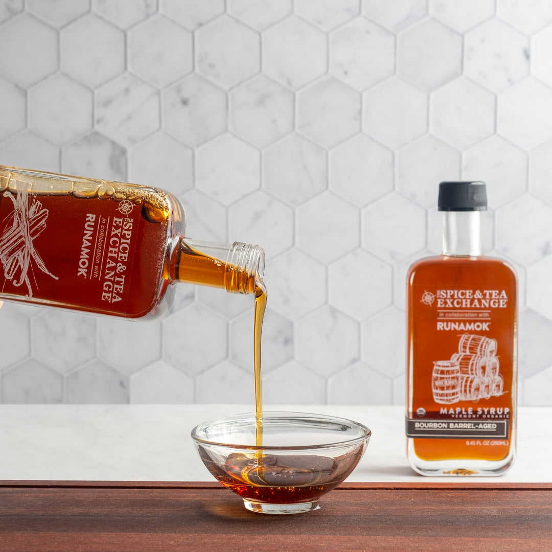 Organic Barrel-Aged Bourbon Maple Syrup is poured into a glass bowl, with another bottle in the background on a wooden surface. White hexagonal tiles create a clean backdrop.