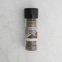 A clear grinder filled with Bourbon Smoked Black Pepper sits upright on a light marble surface.