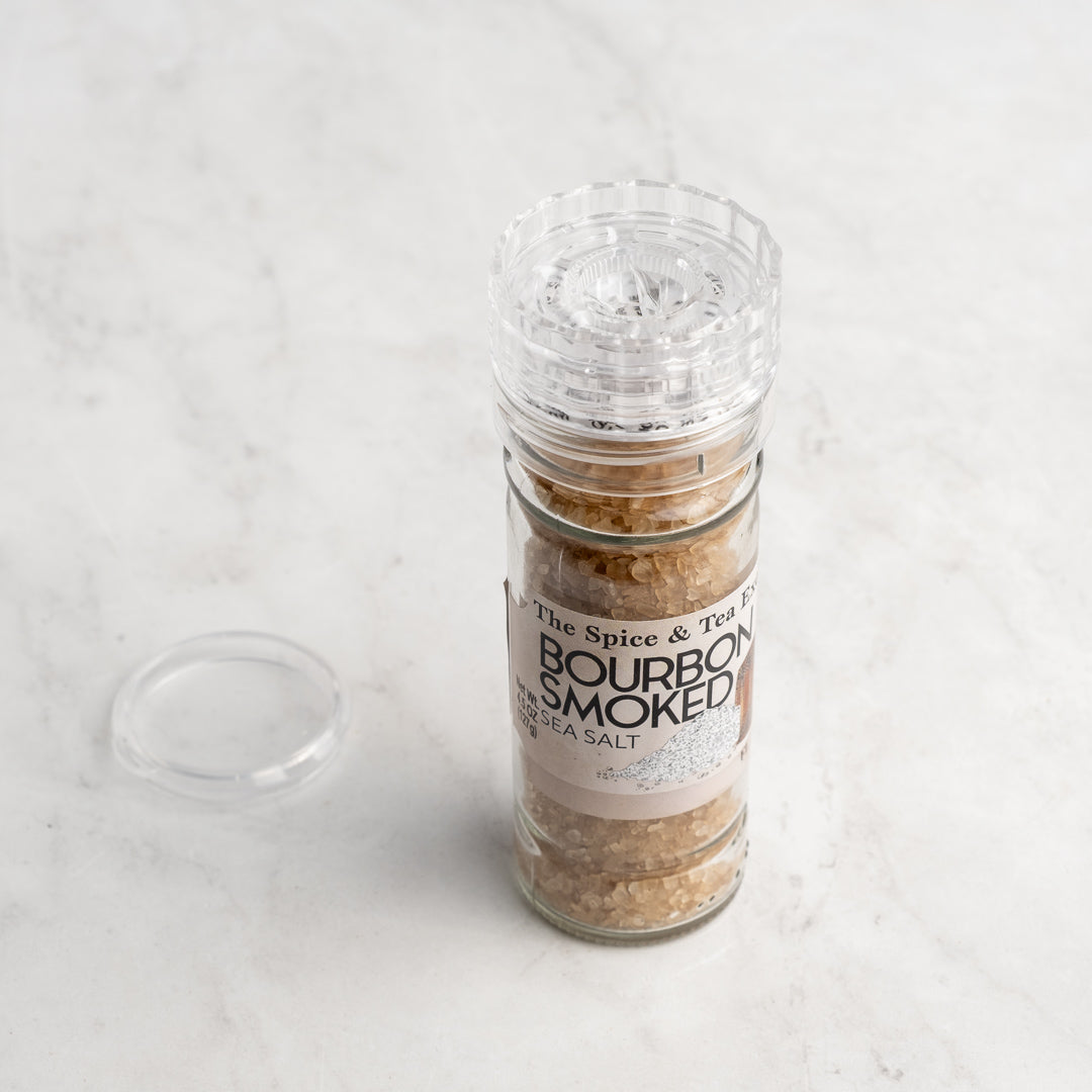 A clear glass grinder of Bourbon Smoked Sea Salt sits on a light marble surface with its plastic lid removed and beside it. Perfect as a finishing salt, the label shows the name: Bourbon Smoked Sea Salt.