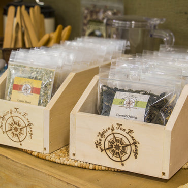 Loose tea leaves in clear plastic bags labeled Coastal Blend and Coconut Oolong are displayed in Large Branded Wood Storage Boxes. The boxes feature The Spice & Tea Exchange branding, with utensils and spice storage items seen nearby.