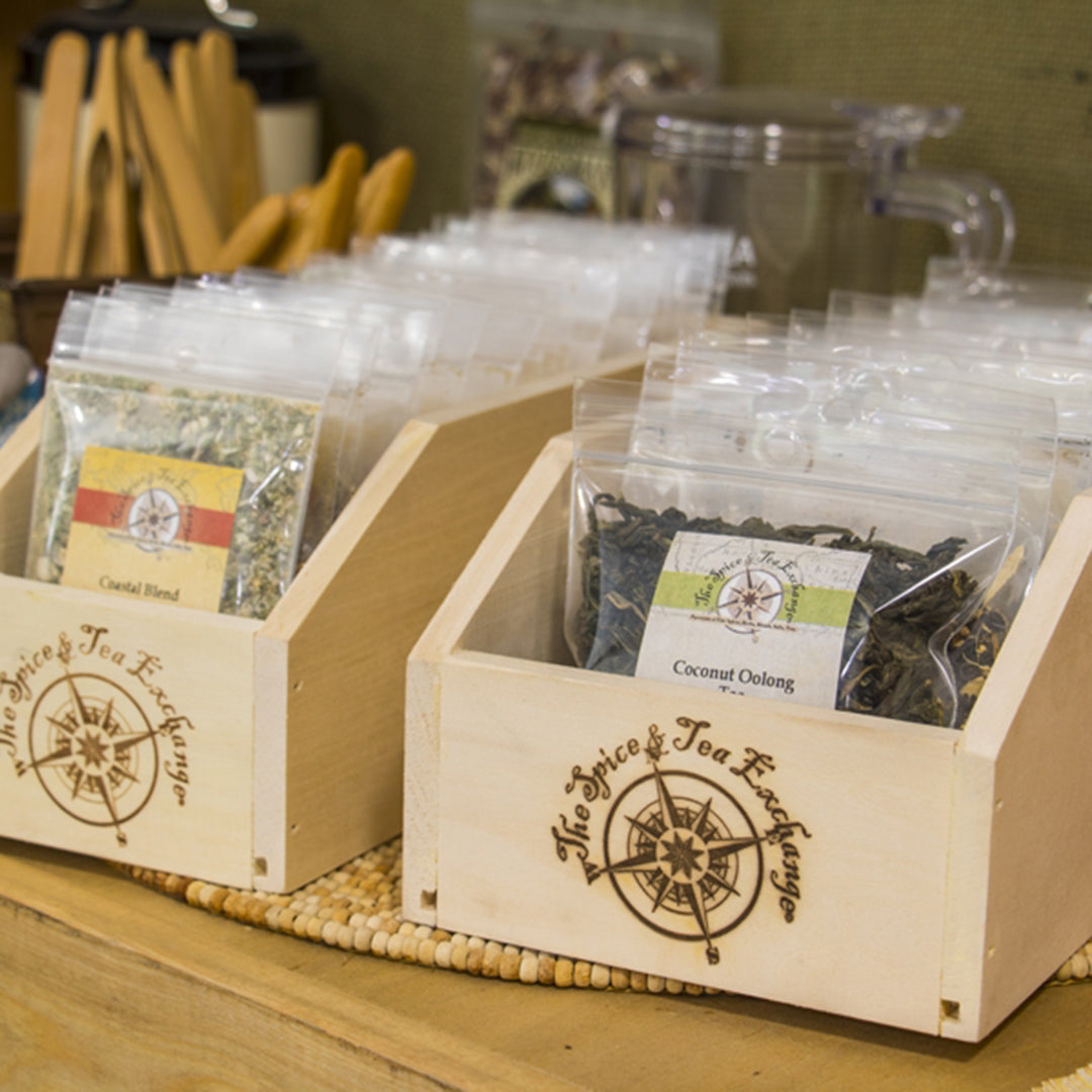 At The Spice & Tea Exchange, clear packets of loose tea blends—including "Coastal Blend" and "Coconut Oolong"—are displayed in a Small Branded Wood Storage Box. Wooden utensils and a clear pitcher can be seen in the background.