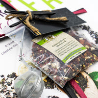 The Cancer Hates Tea Book & Bundle - Ginseng Hippie is shown with a clear bag of herbal tea, metal tea infuser, and gift card, arranged on a vibrant food and tea magazine—ideal for tea lovers and wellness enthusiasts.